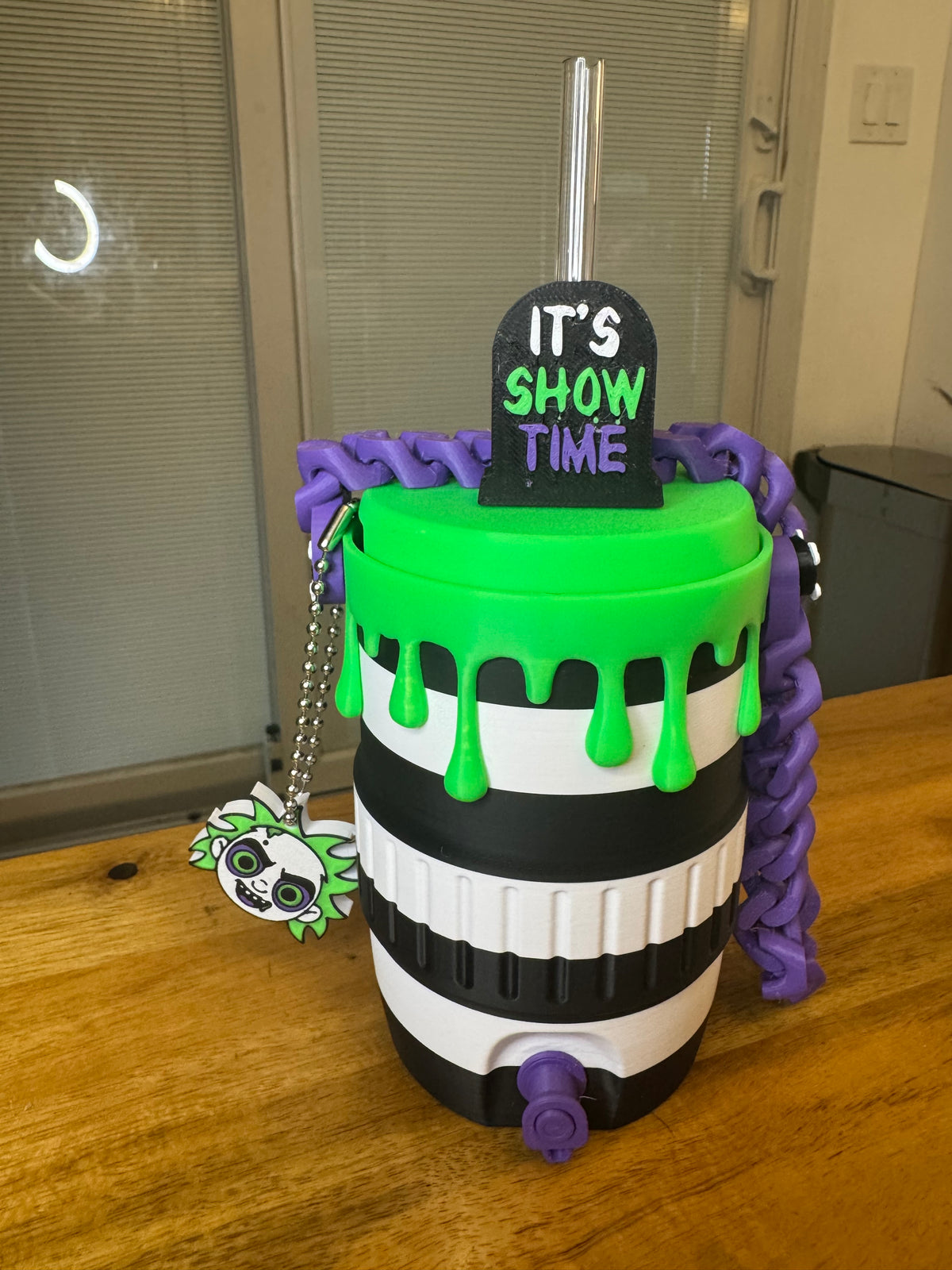 A Halloween-themed spill-proof tumbler cup with a black and white striped body, bright green dripping lid, purple handle, and a tombstone sign reading 'IT'S SHOW TIME' on a wooden table.