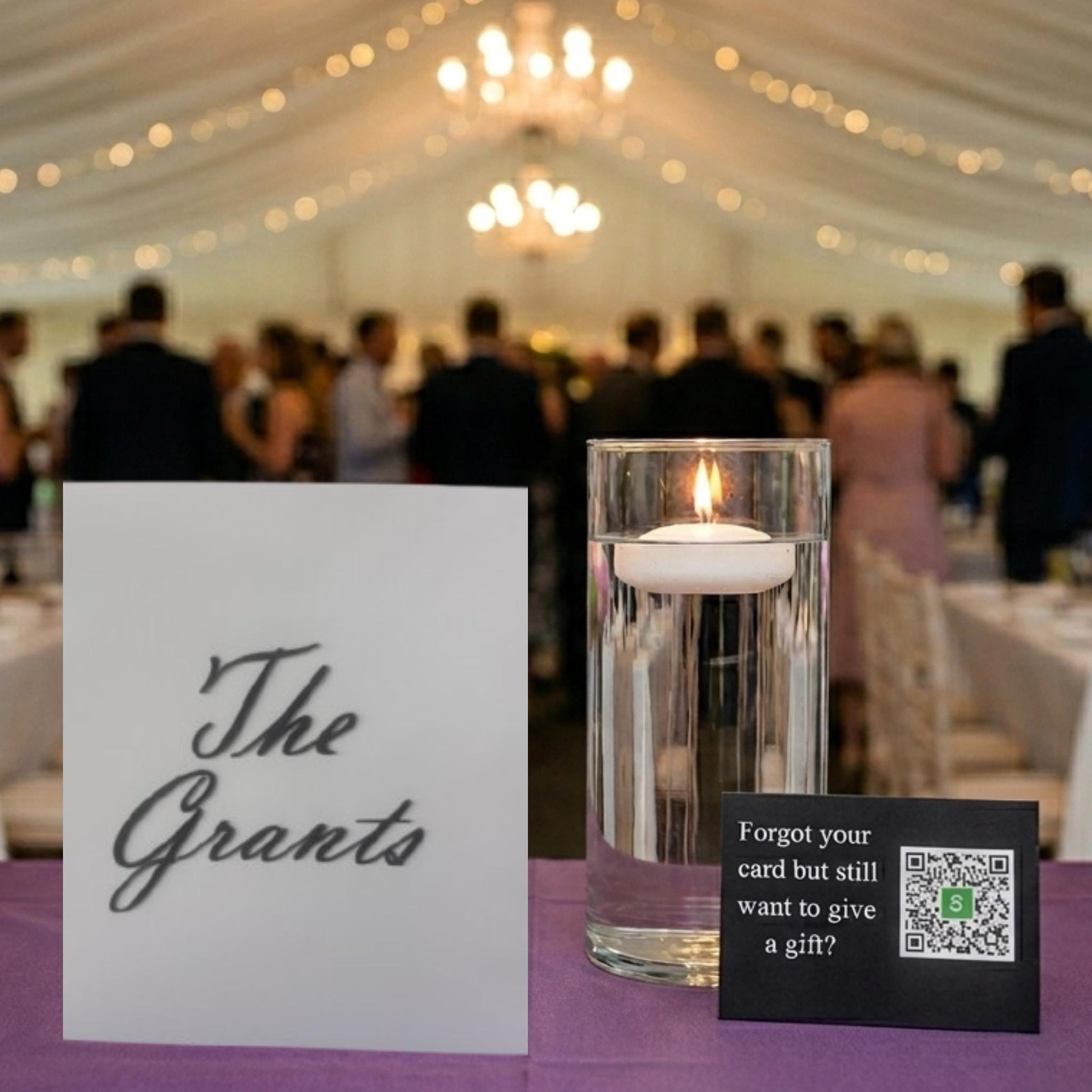 May include: A white rectangular sign with the black text The Grants, a clear cylindrical glass vase containing water and a white floating candle, and a small black rectangular sign with white text and a QR code for gift giving.