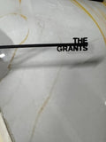 May include: A black metal sign featuring a thin horizontal bar with the text THE GRANTS in a bold, sans-serif font attached to the right side. The letters are connected to the bar, creating a single, unified piece of custom signage.