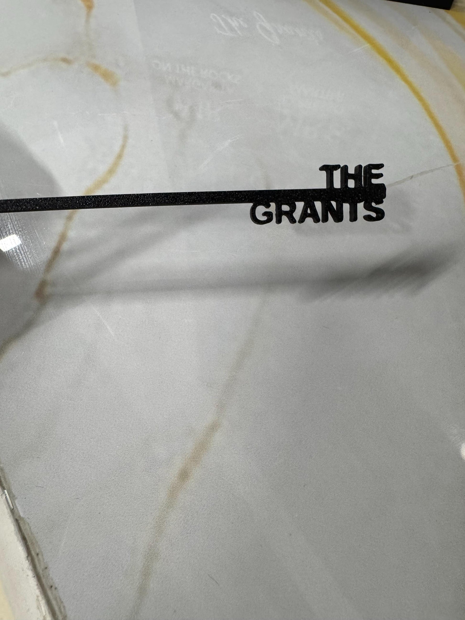 May include: A black metal sign featuring a thin horizontal bar with the text THE GRANTS in a bold, sans-serif font attached to the right side. The letters are connected to the bar, creating a single, unified piece of custom signage.