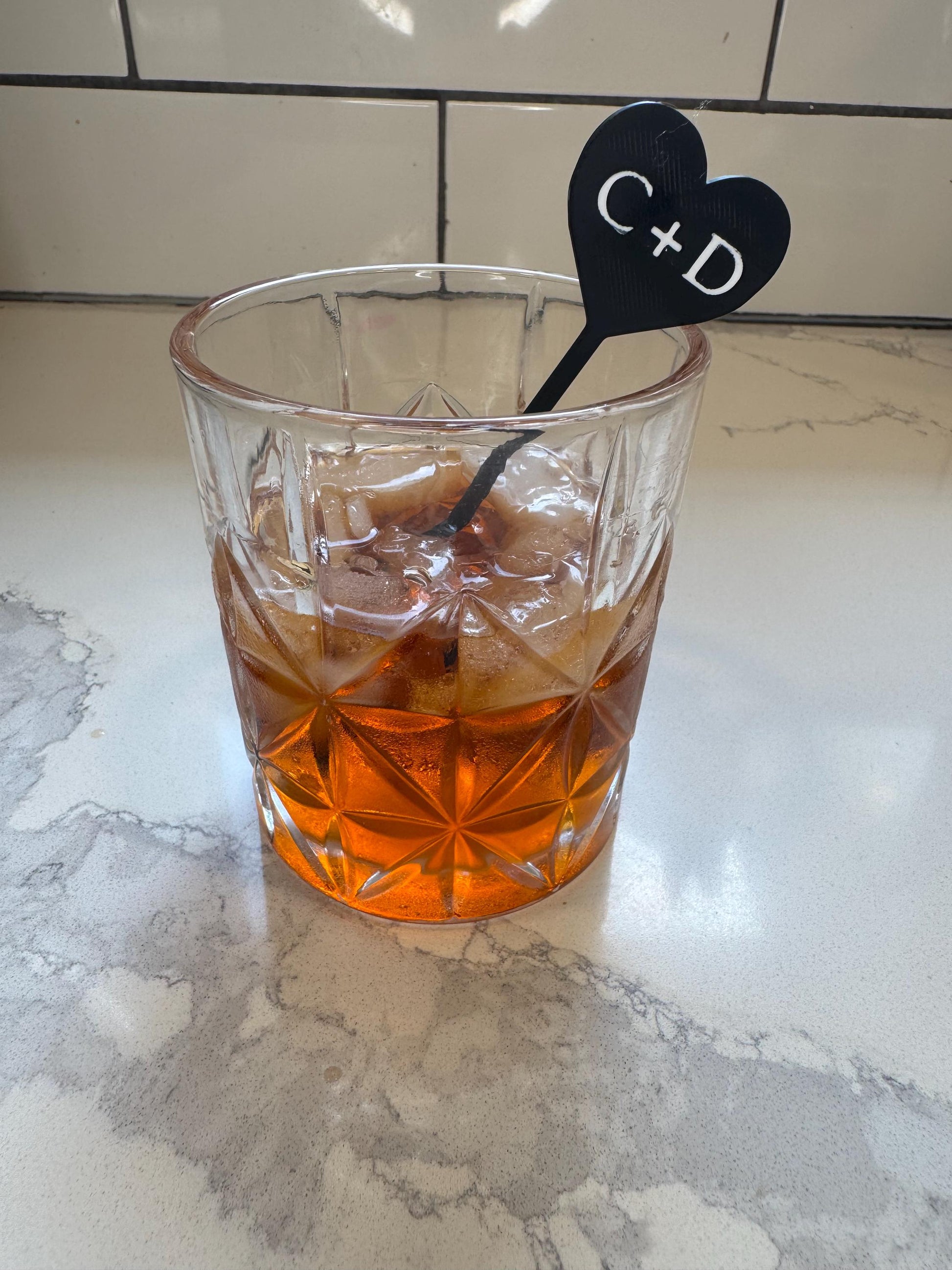 May include: A clear glass tumbler with a geometric diamond pattern filled with an amber-coloured liquid and ice cubes. A black heart-shaped drink stirrer with the white text C+D is inserted into the glass, resting against the rim.