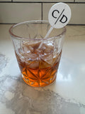 May include: A clear, faceted glass tumbler filled with amber liquid and ice cubes, featuring a white circular drink stirrer with a thin vertical line separating the black letters C and D, positioned upright inside the glass.