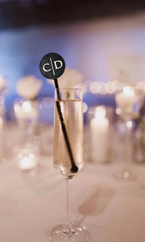 May include: A tall, clear glass flute filled with a light-coloured liquid, featuring a black circular drink stirrer. The stirrer has a thin stem and a round top displaying the white capital letters C and D separated by a vertical line.