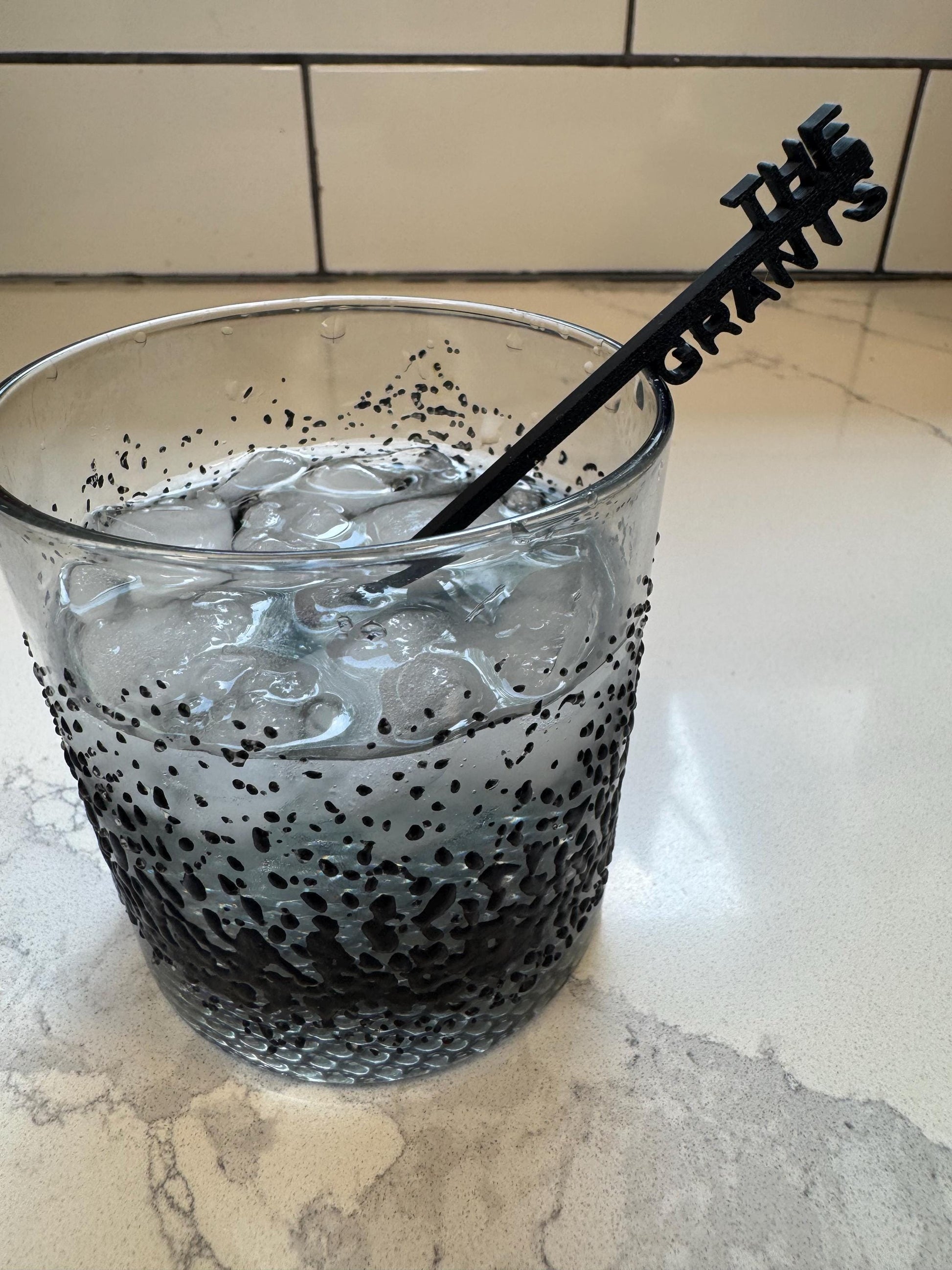 May include: A clear glass tumbler featuring a black speckled gradient pattern filled with ice cubes and a black stirring stick with the text THE GRANTS embossed at the top. The glass has a textured exterior with dense black dots at the base.