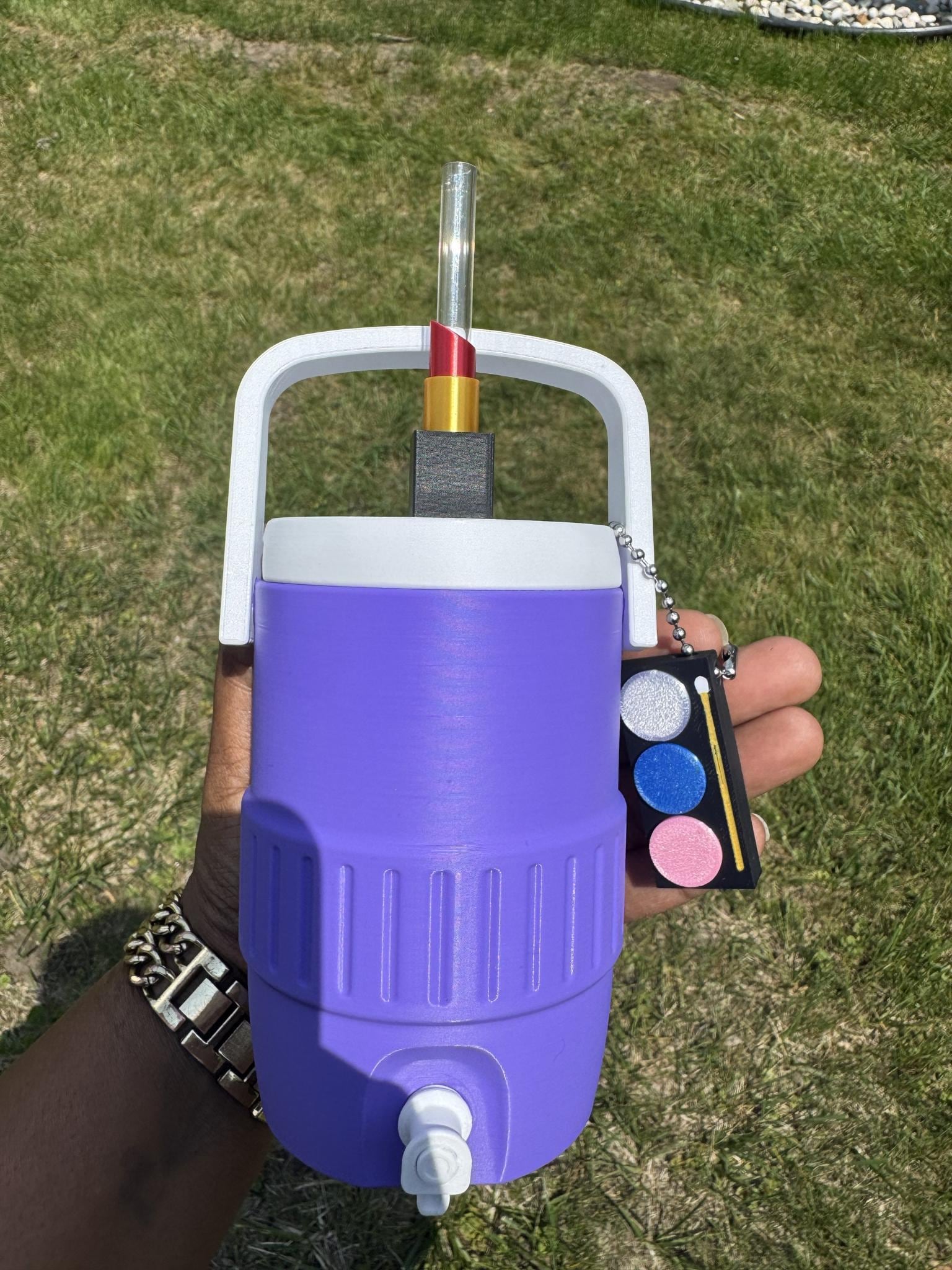 May include: A purple insulated beverage cooler with a white handle and a white spigot at the base. The lid features a clear straw topped with a red and gold lipstick-shaped topper. A small black rectangular makeup palette charm hangs from the handle.